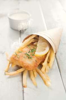 Fish and chips. Fried fish fillet with french fries wrapped by paper cone, on bright wooden background. Fresh cooked with hot smoke.