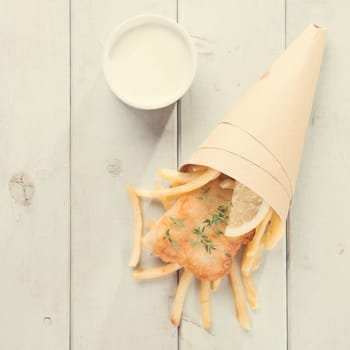 Fish and chips. Top view fried fish fillet with french fries wrapped by paper cone, on bright wooden background in vintage tone.