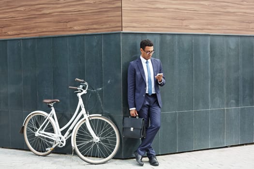 Elegant employee with briefcase using cellphone while standing by corner of modern building with bicycle near by