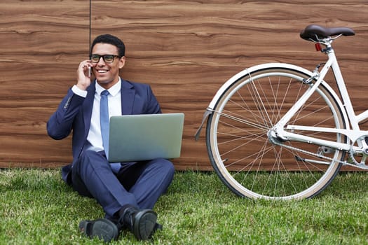Smiling businessman with laptop sitting on lawn and speaking on cellphone