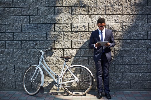 Handsome employee with touchpad networking by wall of modern building with bicycle near by