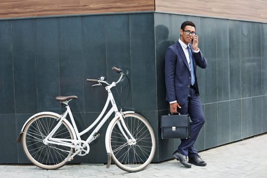 Modern agent speaking on the phone while standing by corner of modern building with bicycle near by