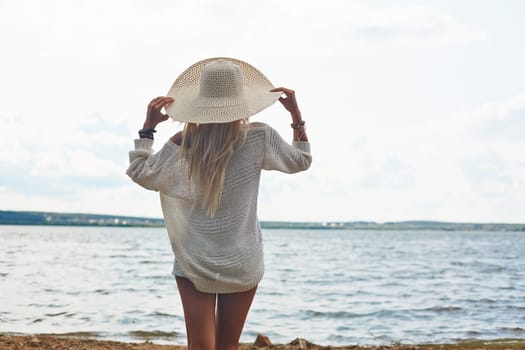 Back view of young woman in hat and white pullover standing by the sea