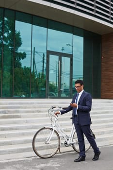 Young businessman with bicycle and cellphone passing by modern building