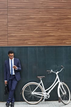 Handsome businessman with briefcase using cellphone while standing by modern building with bicycle near by