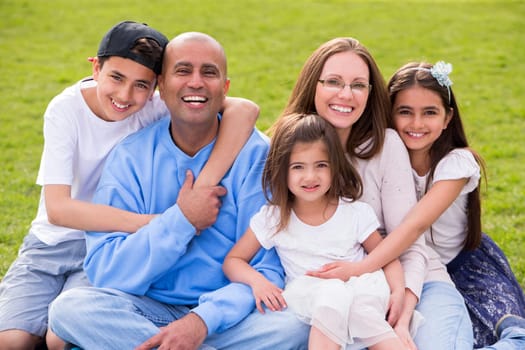 A portrait of a mixed ethnicity family, mum and dad have three cute children between the ages of 4 to 10. They are sitting down and smiling happily at the camera.