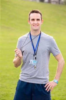 A male sports teacher stands outdoors on a field, he is holding a whistle.