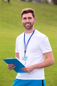A male sports teacher stands outdoors on a field, he is holding a clipboard.