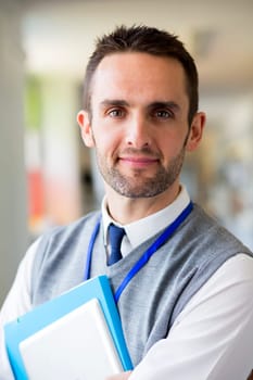 A happy male teacher dressed smartly and smiling in a school corridor. He is holding folders and a digital tablet.