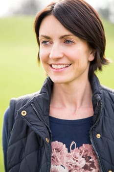 Head and shoulder portrait of a woman looking off into the distance, she is outdoors. A grassy area can be seen behind her, She smiles.
