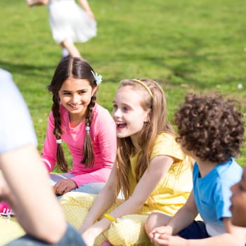 Small group of children sitting on the grass having a lesson outdoors. Only side of the teacher can be seen. The children look to be listening and enjoying themself.