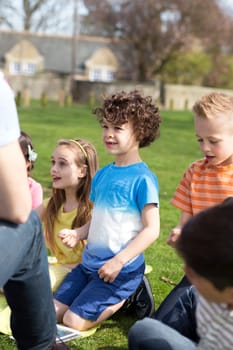 Small group of children sitting on the grass having a lesson outdoors. Only side of the teacher can be seen. The children look to be listening.