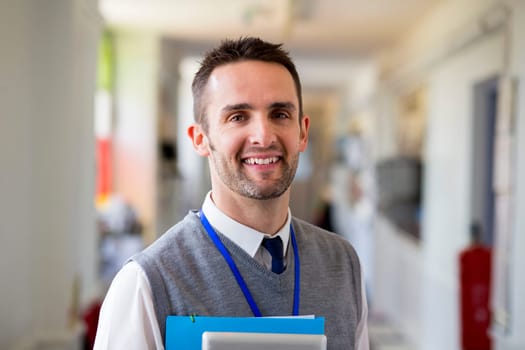 A happy male teacher dressed smartly and smiling in a school corridor. He is holding folders and a digital tablet.