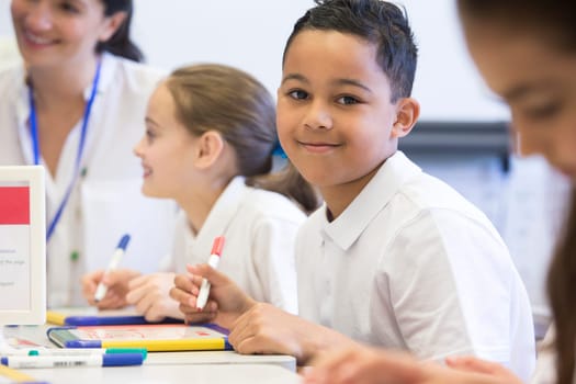 School boy smiles at the camera as he sits at his desk while working.