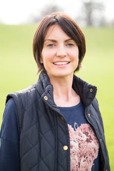 Head and shoulder portrait of a woman looking at the camera, she is outdoors. A grassy area can be seen behind her, She smiles.