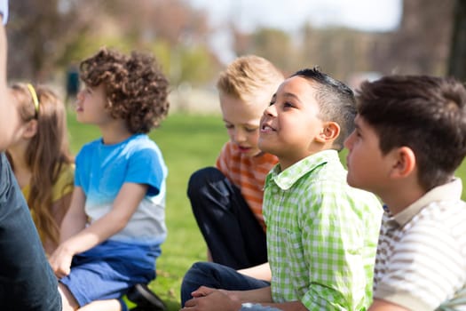Small group of children sitting on the grass having a lesson outdoors. Only side of the teacher can be seen. The children look to be listening.