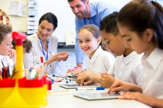 A group of school children can be seen working on digital tablets, two teachers can be seen behind them helping and supervising