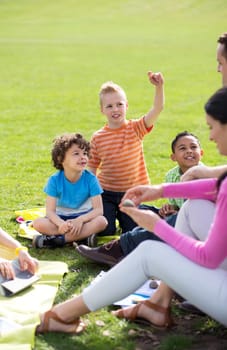 Small group of children sitting on the grass having a lesson outdoors. Only side of the teacher can be seen. The children look to be listening and enjoying themself.
