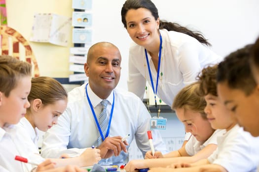 A male and female teacher sit supervising a group of children who are working on whiteboards and digital tablets.