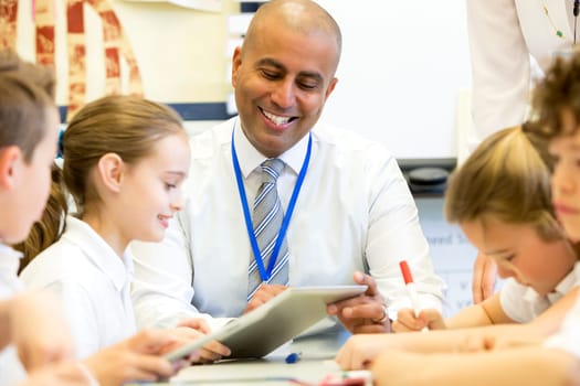 A male teacher sits supervising a group of children who are working on whiteboards and digital tablets.