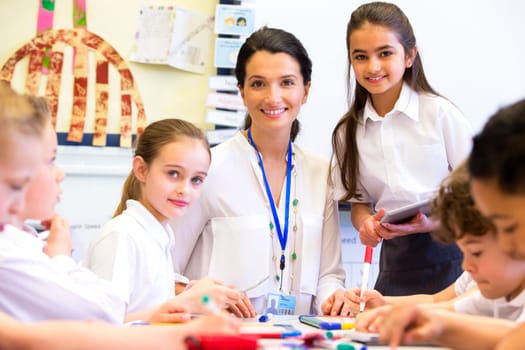 A female teacher and students are looking at the camera smiling. The image has been shot in a school classroom.