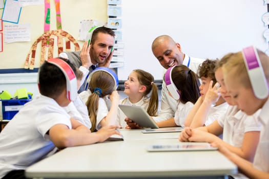 Group of children wearing colourful wireless headsets while working on digital tablets, two teachers can be seen supervising the students in the classroom