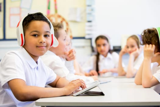 Group of children wearing colourful wireless headsets while working on digital tablets, a little boy can be seen smiling in the foreground looking at camera.