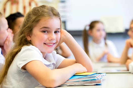 School girl smiles at the camera as she sits at her desk while resting on a big pile of paperwork.