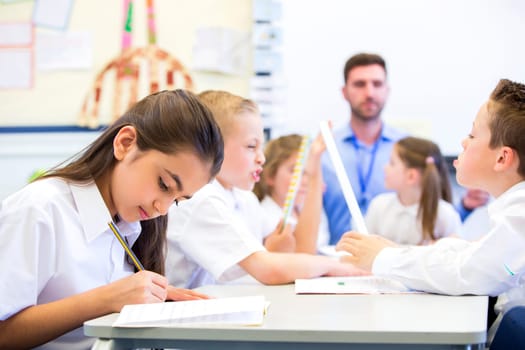 Schoolgirl sitting at a desk with other classmates while working. Her head is down while she concentrates. The teacher can be seen in the background.