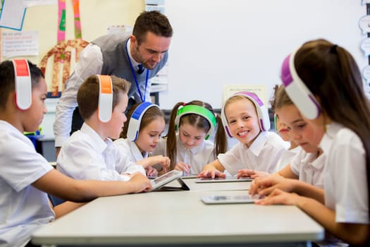 Group of children wearing colourful wireless headsets while working on digital tablets, the teacher can be seen supervising the students in the classroom
