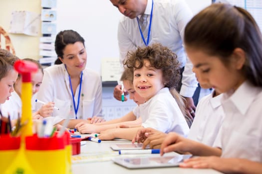A group of school children can be seen working on digital tablets, two teachers can be seen behind them helping and supervising