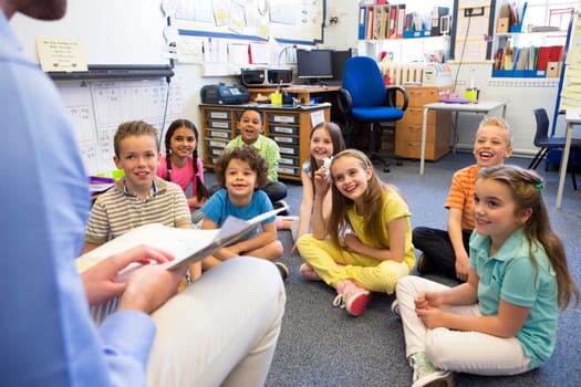 A group of children sit on the floor cross legged, listening to the teacher read a story.
