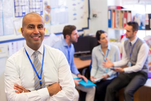 School teachers gather in a small school office for a chat. They look happy. The focus is on the man who stands in the foreground with the others sitting out of focus behind. The man smiles at the camera.
