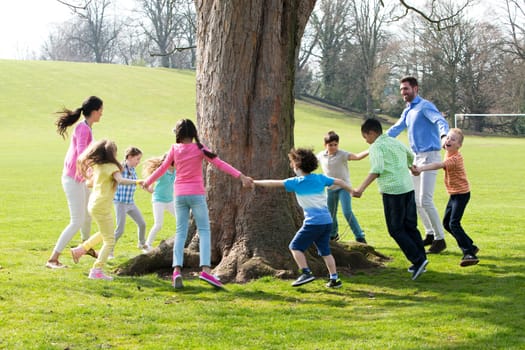 A group of children with two adults holding hands and dance around the trunk of a tree.