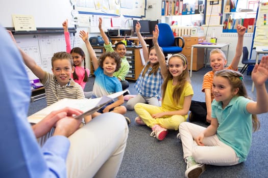 A group of children sit on the floor cross legged, listening to the teacher. They all have their hands raised in the air to answer a question.