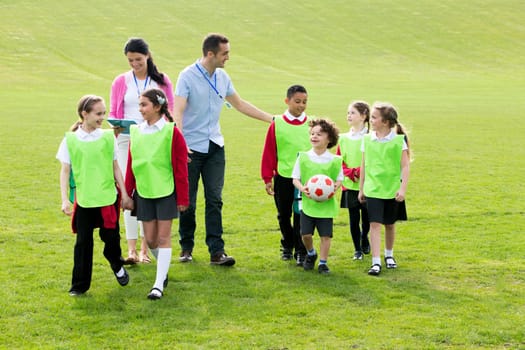 Group of school children wearing bibs and school uniform outside on the grass with their teacher. One of the boys is holding a soccor ball.