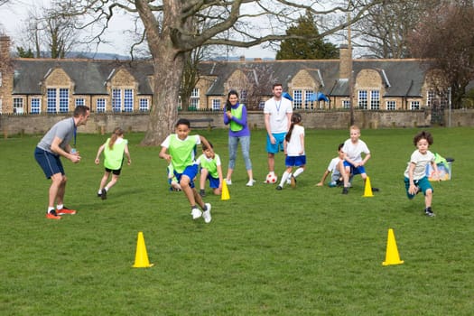 Adults on grassed area with school children supervising a football training session, Everyone can be seen running around cones. School building can be seen in the background.