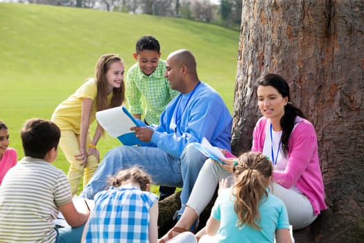 Small group of children sitting on the grass having a lesson outdoors. Male and female teacher can be seen. The children look to be listening and enjoying themself.