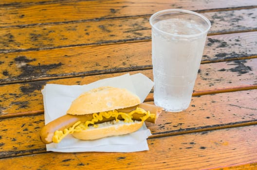 Bockwurst with mustard in the bun and a cup of mineral water on a brown wooden table on a white napkin.