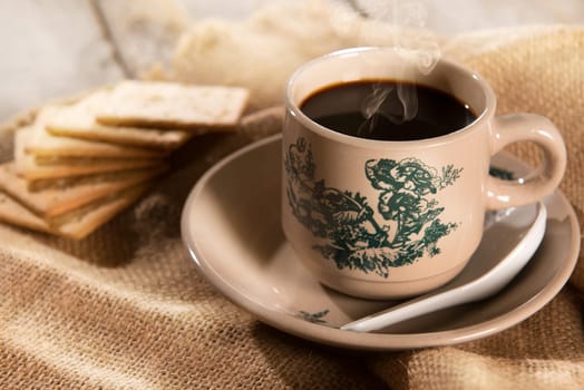 Steaming traditional oriental Chinese kopitiam style dark coffee in vintage mug and saucer with soda crackers. Fractal on the cup is generic print. Soft focus setting with dramatic ambient light on dark wooden background.