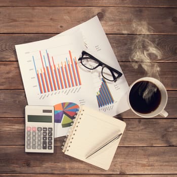 Top view workspace with booklet, pen, glasses, cup of coffee and graphs. Wooden table background in vintage toned.