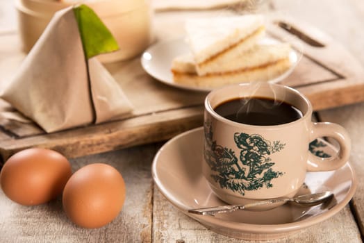Traditional kopitiam style Malaysian coffee in vintage mug and saucer and breakfast with morning sunlight. Fractal on the cup is generic print. Soft focus dramatic ambient light over wood table.