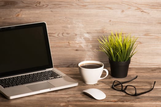 Wooden workplace with laptop, cup of hot coffee, mouse, glasses and pot plant, in dramatic light vintage toned.
