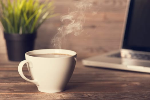 Morning caffeine concept. Wooden business desk with laptop, cup of hot coffee and pot plant, in dramatic light vintage toned.