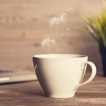 Cup of hot steaming coffee on working table, laptop and plant pot at background. Wooden desk in dramatic light vintage toned.
