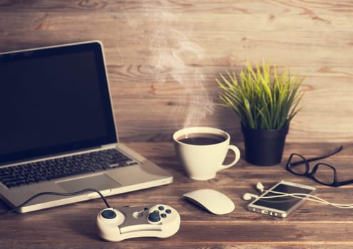 Wooden office workplace, table with laptop, online games controller, cup of hot coffee, mouse, glasses, smartphone, earphones and pot plant, in dramatic light vintage toned.