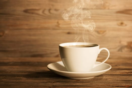 Steaming coffee in white mug and saucer in soft focus setting with dramatic ambient light, over dark wooden background.