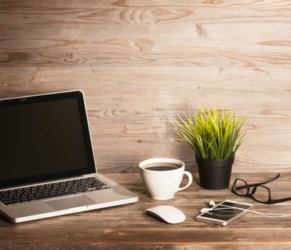Wooden office interior, table with laptop, cup of hot coffee, mouse, glasses, smartphone, earphones and pot plant, in dramatic light vintage toned.