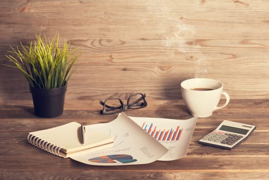 Workplace with booklet, pen, glasses, cup of coffee and graphs. Wooden table background in vintage toned.