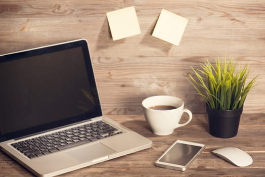 Wooden working desk with laptop, cup of hot coffee, mouse, smartphone and pot plant, in vintage toned.